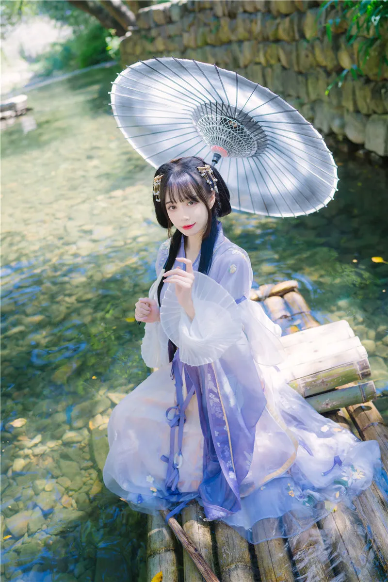 浅紫色汉服少女跪坐在清澈溪流上的竹筏边缘,一手撑银色油纸伞,一手微抬,阳光穿透树叶在水面与白纱袖口留下光斑