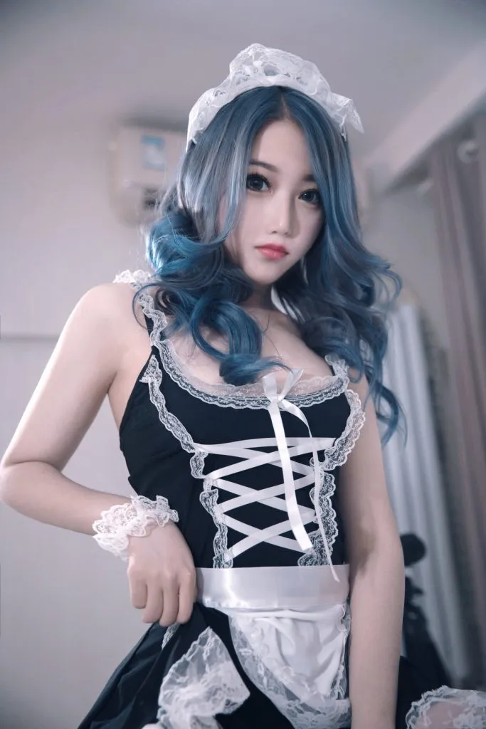 Eloise软软在冷调室内,蓝绿色大波浪卷发女子头戴白色蕾丝女仆发带,身穿黑白撞色的蕾丝边女仆吊带裙,正面看向镜头并用手轻拉裙摆