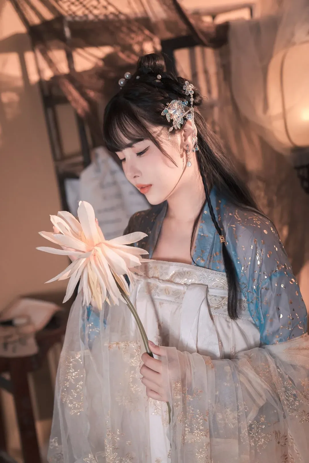 恋恋艾妮穿浅色汉服站在暖光室内，低头捧着大朵花枝的近景造型