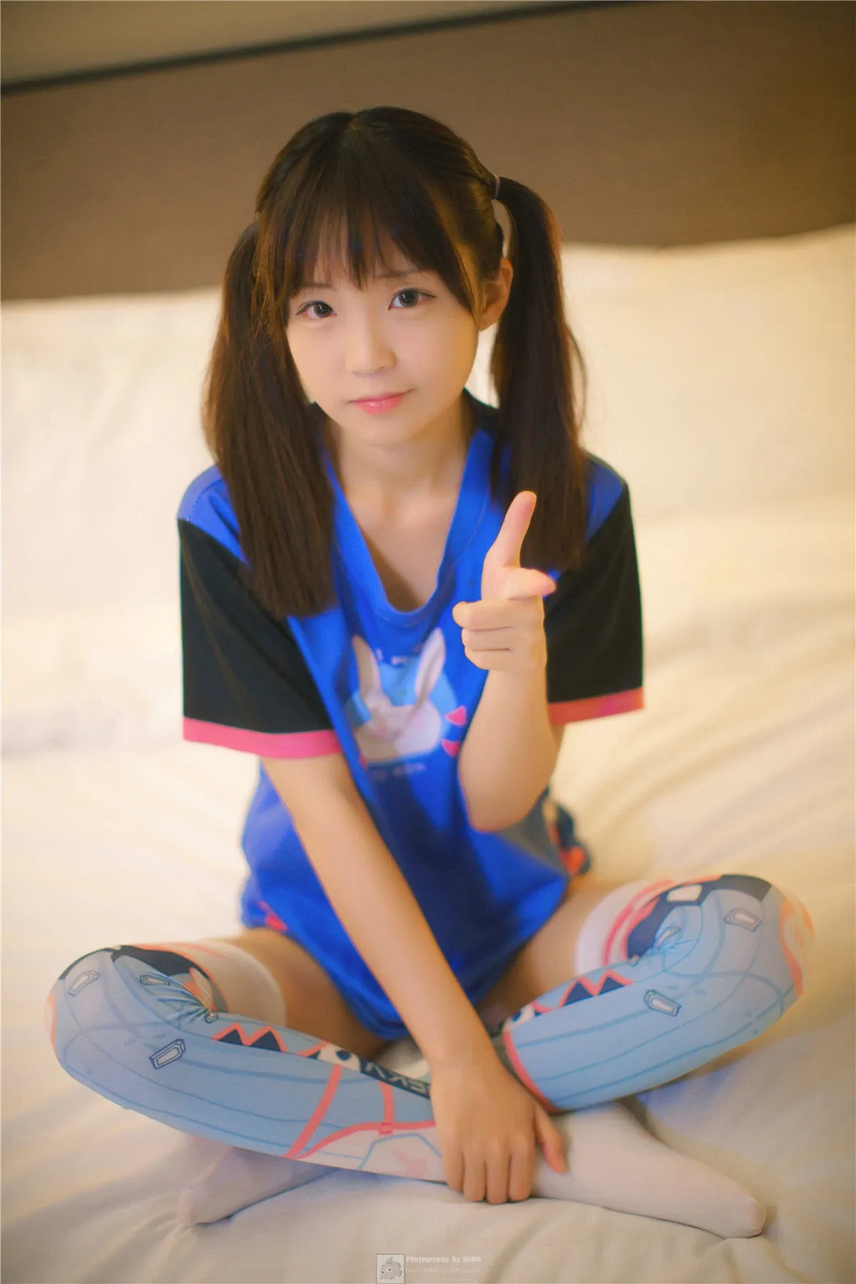 御子Yumiko坐在床上穿蓝黑配色上衣，双马尾造型对镜头比手势