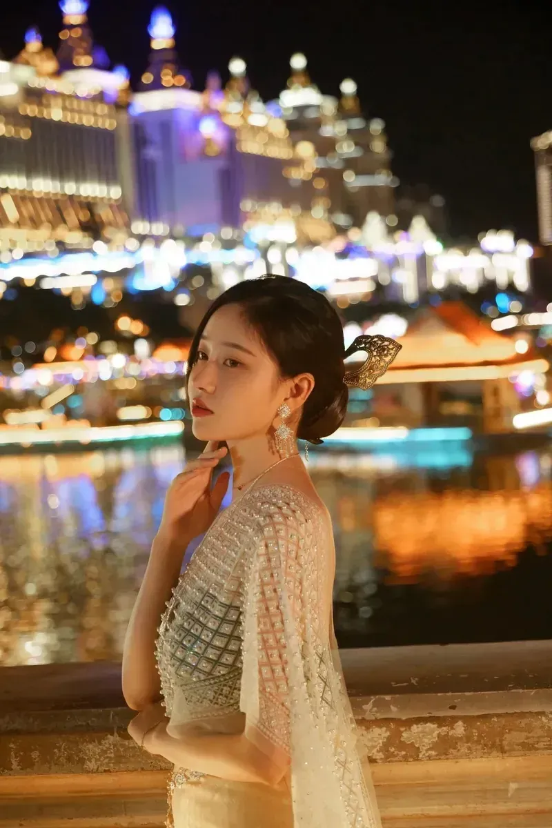 年年nnian盘发佩戴金色发饰,穿白色亮片礼服站在夜景水边栏杆前,背景有城市灯光