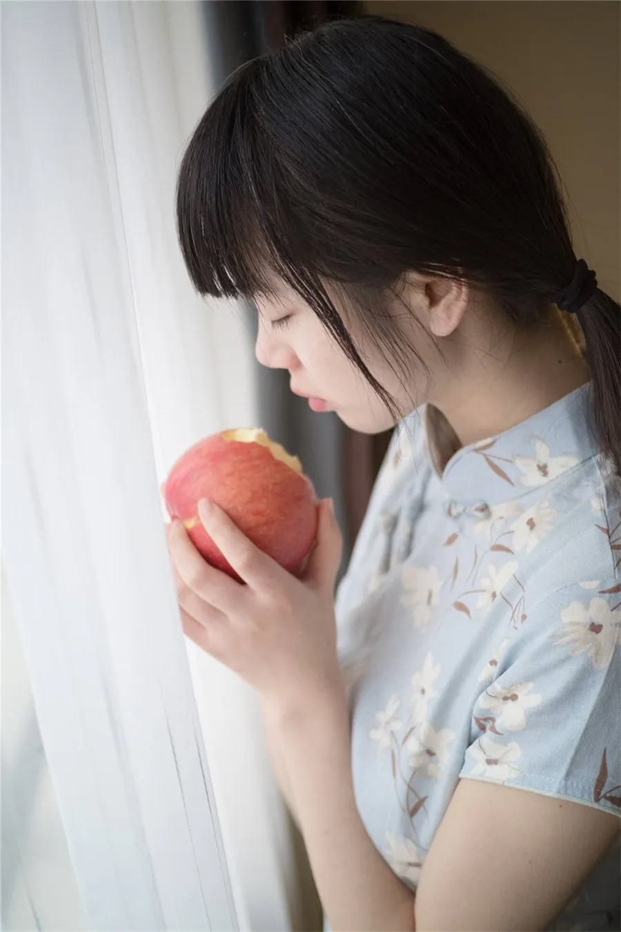 西尔酱低马尾浅蓝花纹旗袍手持红色苹果靠近窗帘的写真